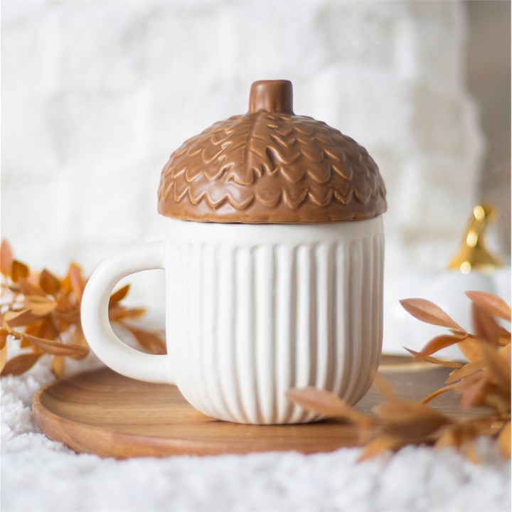 White mug with a brown acorn-shaped lid on a wooden coaster with autumn leaves.